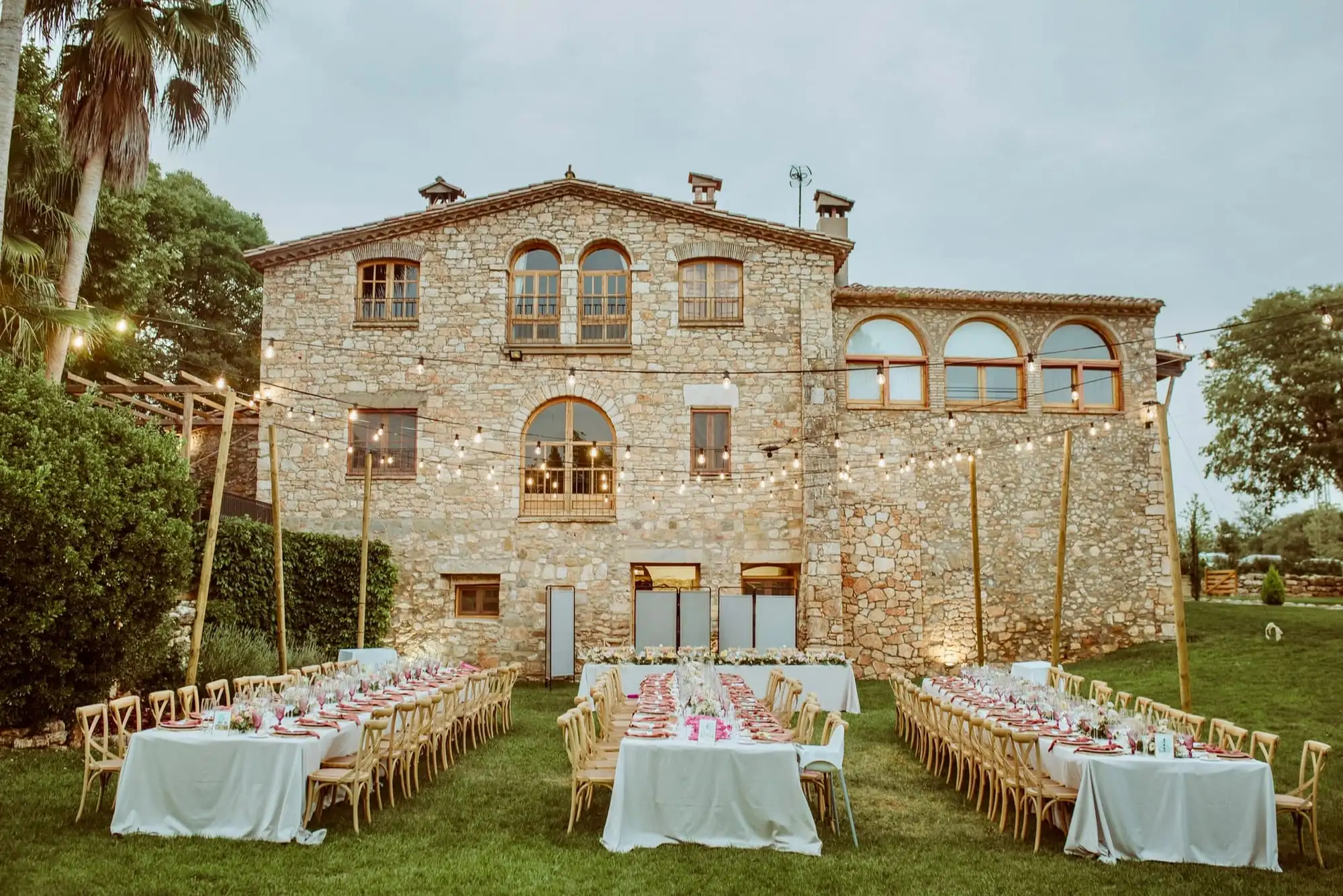 Long banquet tables set on the lawn in front of the stone farmhouse, under strings of lights
